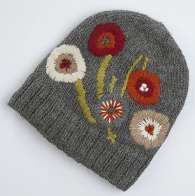 Grau gestrickte Alpaka-Mütze mit bunter, gestickter Blumenverzierung in Rot, Orange, Weiß und Beige, grüne Stängel und Blätter, grobe Strickstruktur, warme Winterkopfbedeckung