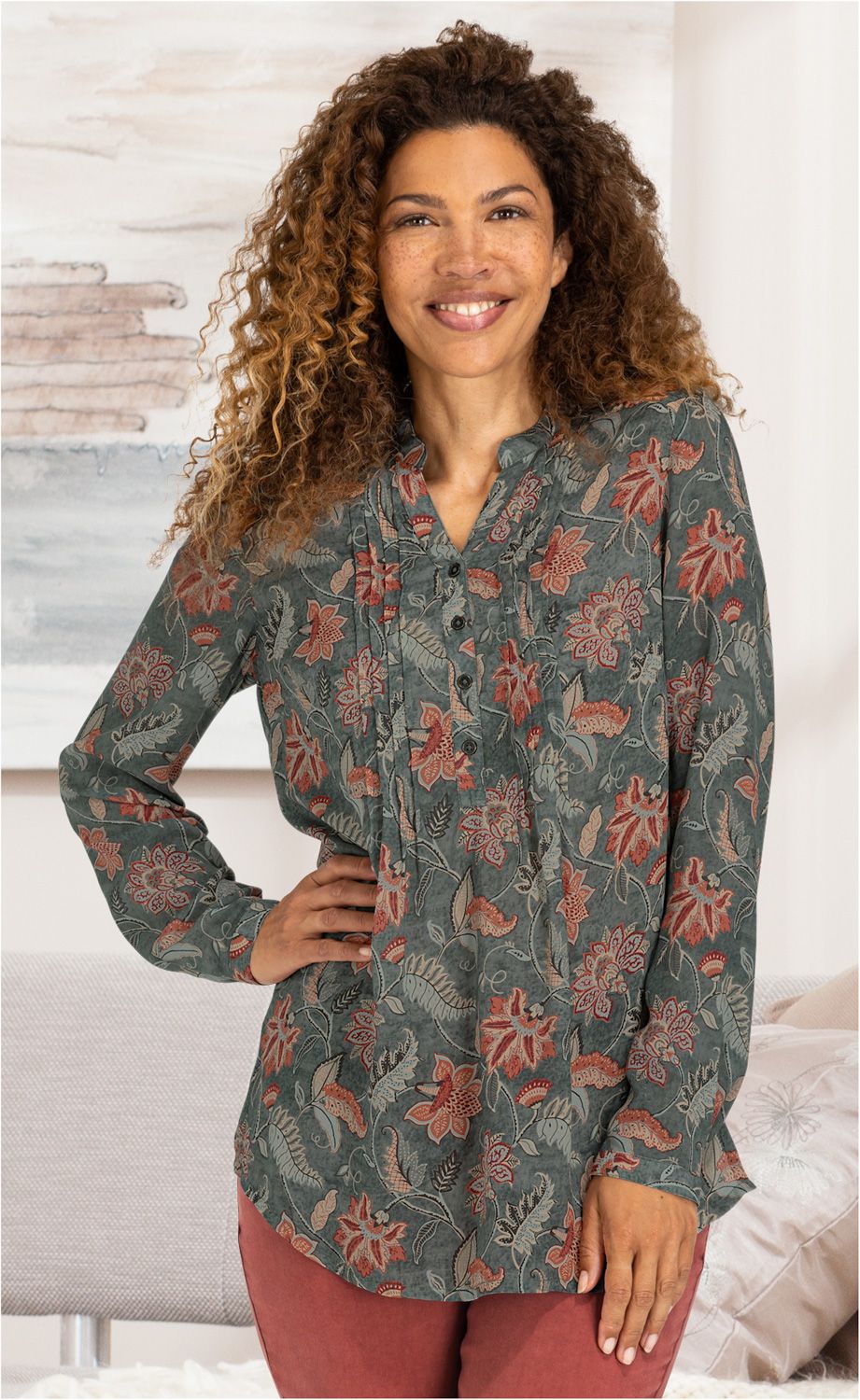 Frau mit lockigen braunen Haaren trägt eine grüne Modal-Bluse mit roten und beigen Blumenmustern, Knopfleiste und Stehkragen, kombiniert mit roten Hosen, steht lächelnd vor hellem Hintergrund, Hand in Hüfte gestützt.