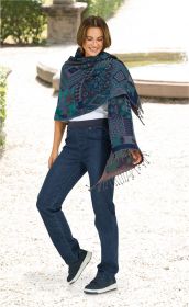 Frau trägt blau-grünen Jacquard-Schal mit geometrischem Muster und Fransen, kombiniert mit weißem Oberteil, blauen Jeans und schwarzen Sneakern, steht lächelnd im hellen Garten mit Springbrunnen im Hintergrund