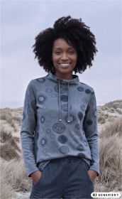 Frau mit lockigem Haar trägt blaues Baumwoll-Shirt mit langen Ärmeln und Muster aus kreisförmigen Motiven, Hände in Hosentaschen, steht in sandiger Dünenlandschaft bei bewölktem Himmel