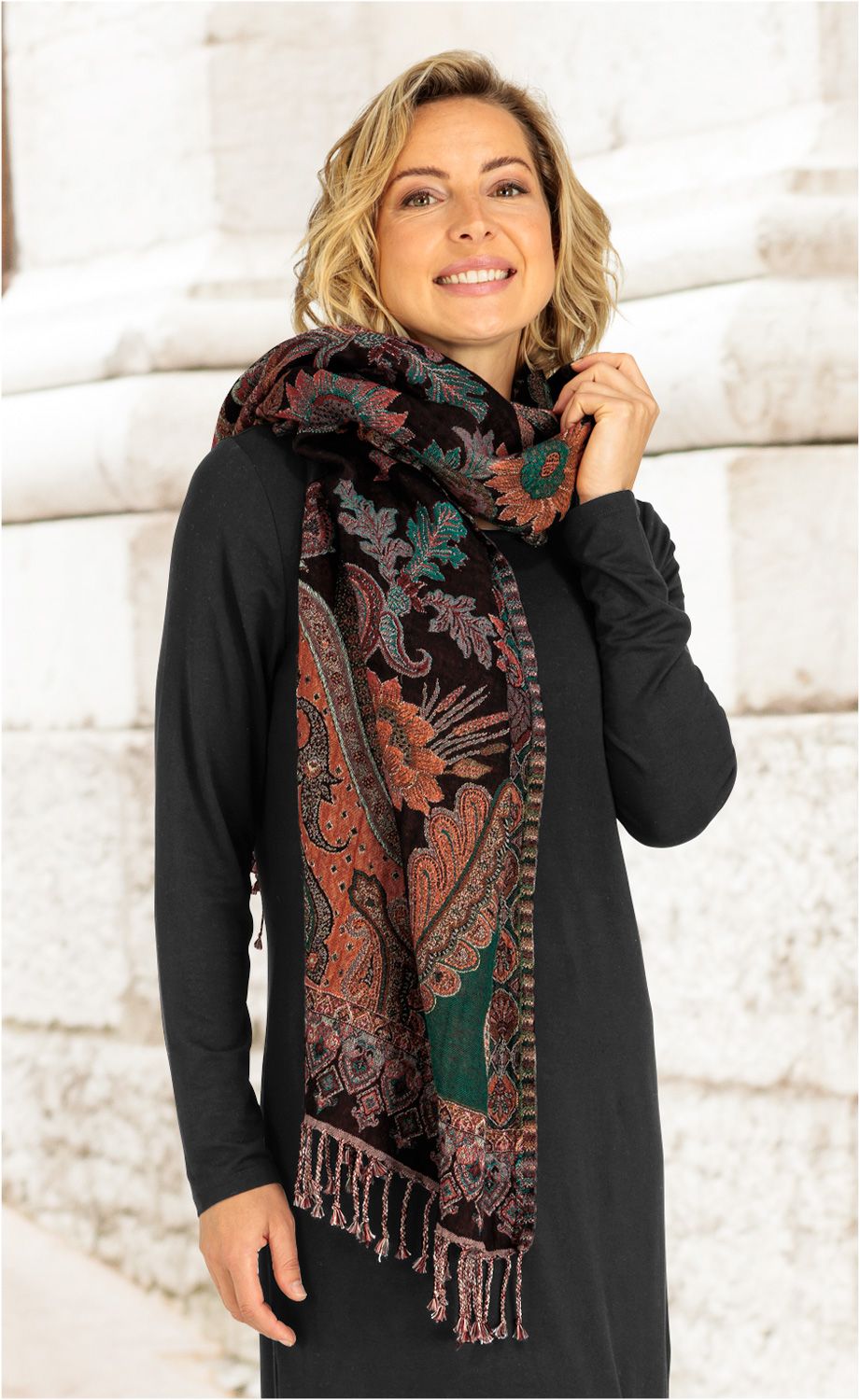 Frau trägt langen Jacquard-Schal mit floralem Muster in Braun, Orange und Grün, der locker um den Hals drapiert ist, vor hellem Mauerhintergrund, schwarzes Kleid, lächelnd, rechte Hand hält Schal am Hals