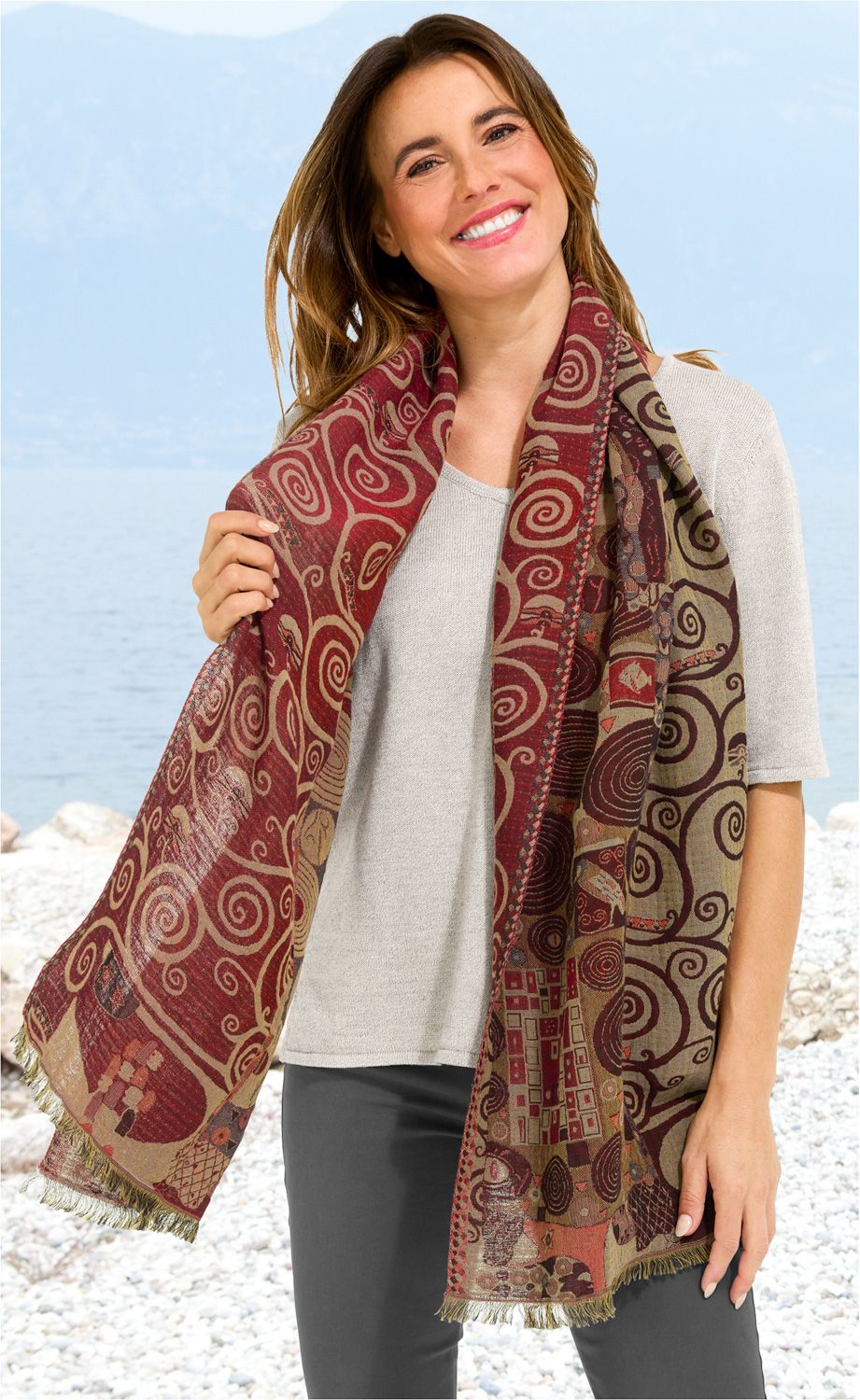 Frau trägt Baumwoll-Mix-Schal 50x180 mit rot-braunem, kunstvollem Muster aus Spiralen und geometrischen Formen, Baumwollmix, lächelnd, heller Pullover, dunkle Hose, Strand mit Kieselsteinen und ruhiges Wasser im Hintergrund