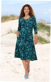 Frau mit lockigen Haaren trägt ein knieumspielendes Viskose-Kleid mit 3/4 Ärmeln und floralen, bunten Mustern, geht barfuß am Strand mit Sanddünen und Gras im Hintergrund, lächelnd, seitliche Taschen vorhanden