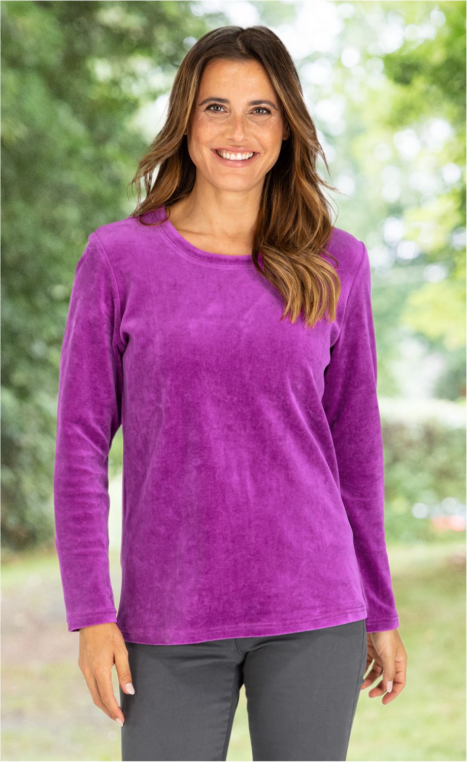 Frau mit langen braunen Haaren trägt violettes Baumwoll-Shirt mit Rundhalsausschnitt und langen Ärmeln, lächelt, steht vor verschwommenem grünen Hintergrund im Freien