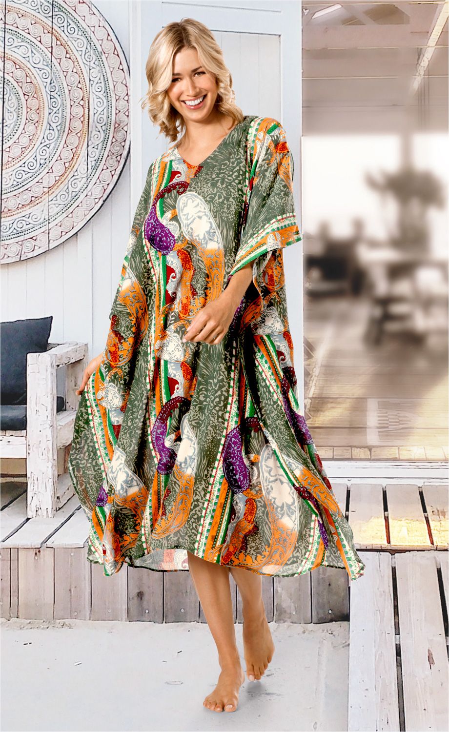 Frau trägt bunten Viskose-Kaftan mit Paisley-Muster in Grün, Orange, Lila und Weiß, lächelt, barfuß, stehend auf Holzterrasse mit Holzmöbeln und dekorativer runder Wandplatte im Hintergrund