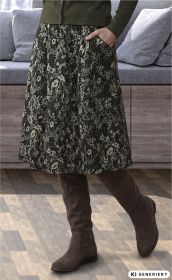 Frau trägt knielangen Rock mit floralem Muster und kniehohe braune Stiefel, Hand in Rocktasche, im Hintergrund graues Sofa mit grauen Kissen, Holzboden