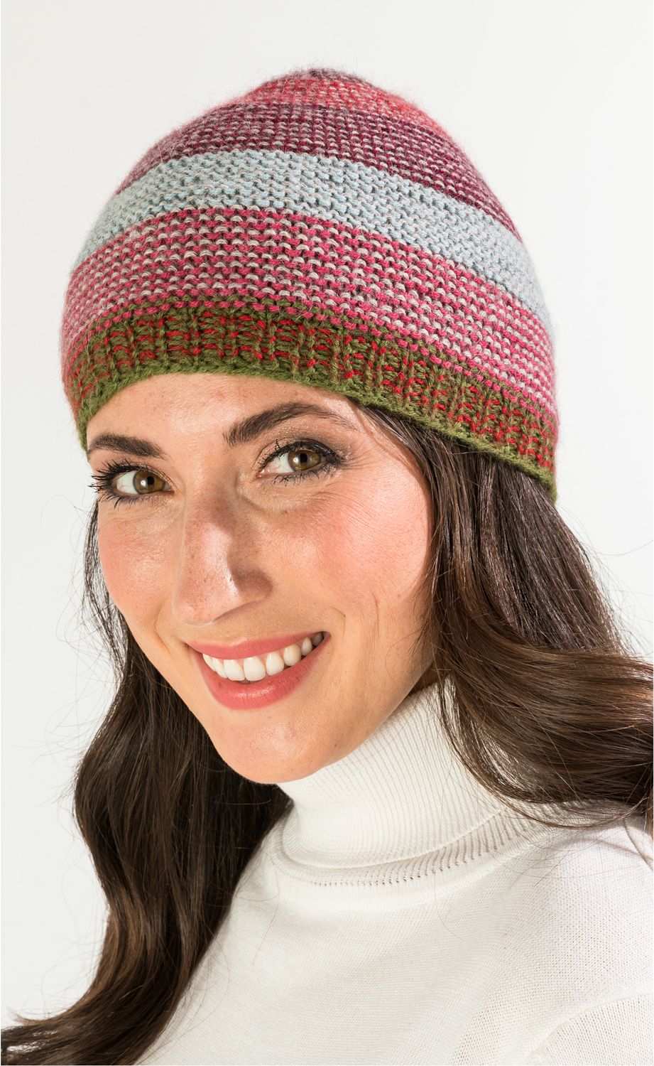 Frau mit langen braunen Haaren und weißem Rollkragenpullover trägt eine mehrfarbige, gestrickte Alpaka-Mütze in Rot-, Rosa-, Hellblau- und Grüntönen, lächelt freundlich vor hellem Hintergrund