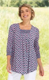 Frau trägt Viskose-Bluse mit blau-pinkem geometrischem Muster, gerafftem Rundhalsausschnitt und 3/4-Ärmeln, lächelt, steht vor grüner Wiese, kombiniert mit weißen Hosen.