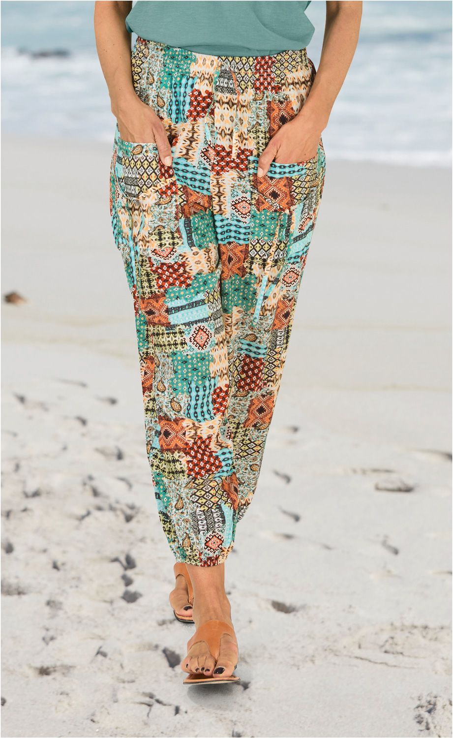 Person trägt bunte Viskose-Hose mit Patchwork-Muster in Türkis, Rot, Braun und Beige, Hände in Taschen, steht barfuß mit braunen Sandalen auf hellem Sandstrand