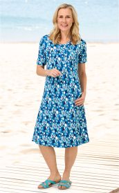 Frau am Strand trägt blaues Viskose-Mix-Kleid mit weißem und türkisfarbenem Blumenmuster, halbarm, knielang, kombiniert mit türkisfarbenen Sandalen, lachend, auf Holzsteg stehend vor Strand und Meer im Hintergrund