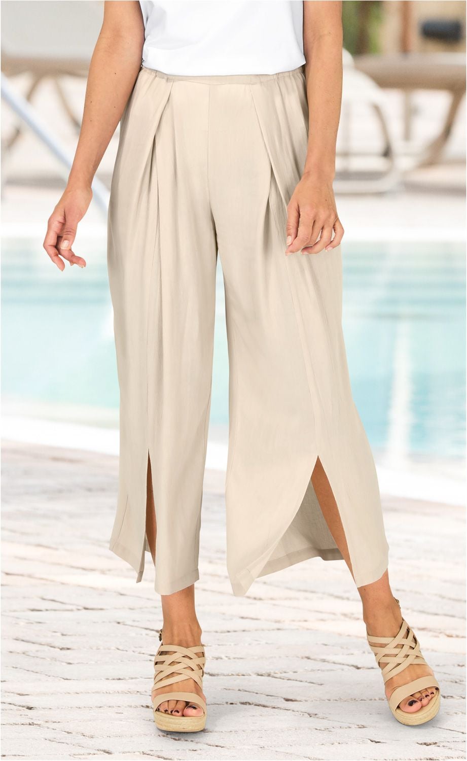 Beige Viskose-Hose mit weitem Bein und seitlichen Schlitzen, getragen von einer Person mit weißen Sandalen, kombiniert mit weißem Oberteil, vor hellem, outdoor Hintergrund mit Pool und gepflastertem Boden