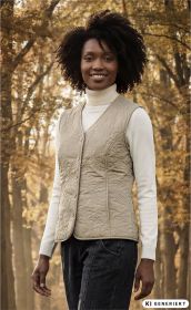 Frau trägt beige gesteppte Baumwoll-Weste mit V-Ausschnitt und Knöpfen, weißem Rollkragenpullover und dunkler Jeans, herbstlicher Wald im Hintergrund mit warmem Licht und goldbraunen Blättern