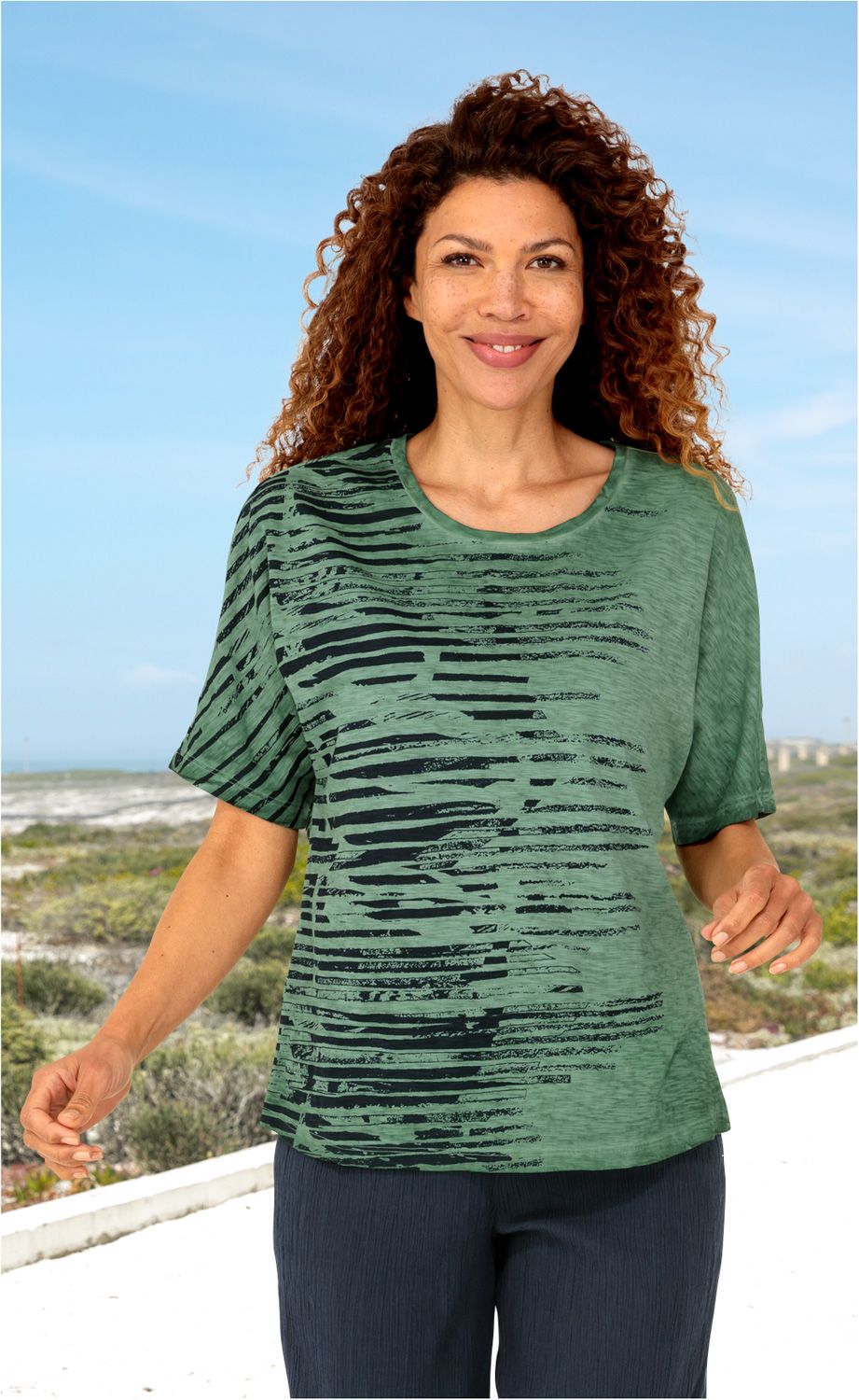 Frau mit lockigen Haaren trägt grünes Baumwoll-Shirt mit schwarzem Streifenmuster, steht draußen bei blauem Himmel und Vegetation, schwarze Hose, lächelnd