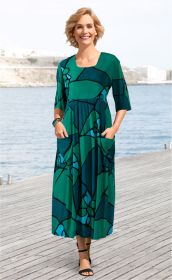 Frau trägt langes Viskose-Kleid in Grün und Blau mit abstraktem Muster, halblange Ärmel, zwei Taschen, schwarze Sandalen, steht auf Holzsteg mit Wasser und Felsen im Hintergrund, lächelt freundlich