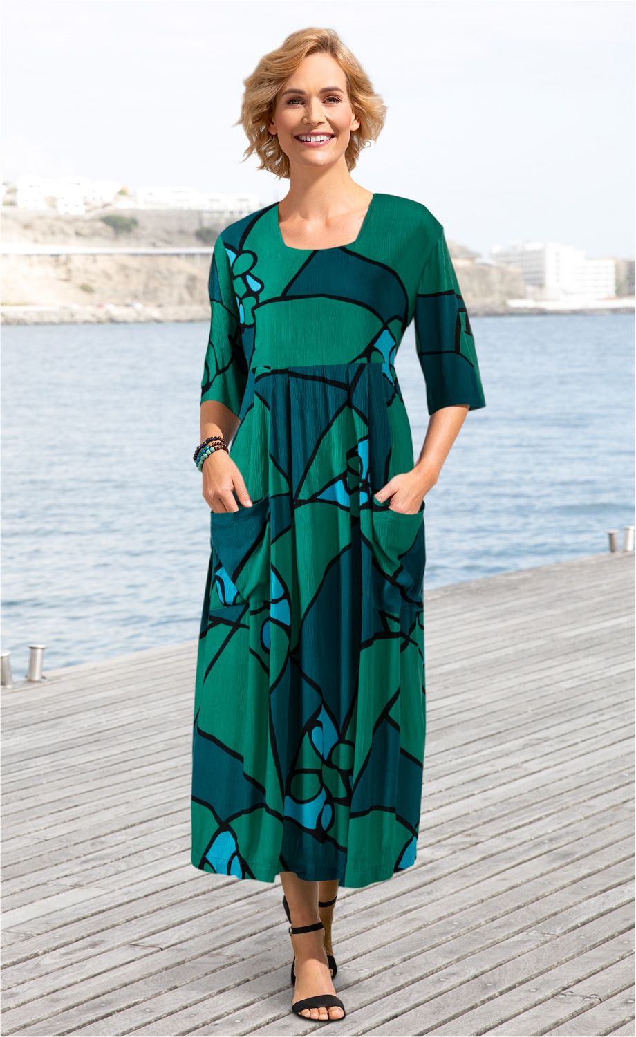 Frau trägt langes Viskose-Kleid in Grün und Blau mit abstraktem Muster, halblange Ärmel, zwei Taschen, schwarze Sandalen, steht auf Holzsteg mit Wasser und Felsen im Hintergrund, lächelt freundlich