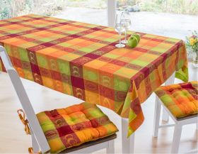 Tisch mit bunter Tischdecke 130x180cm in Orange, Grün und Rot mit Apfel- und Birnenmotiven, zwei weiße Stühle mit passenden Sitzkissen, zwei Gläser mit Wasser und zwei grüne Äpfel auf der Decke, heller Raum mit großer Fensterfront und Gartenblick