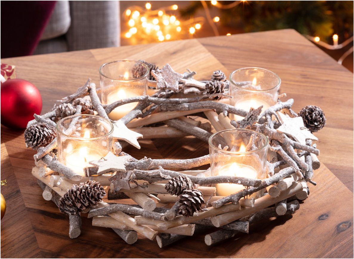 Adventskranz aus Holzstöcken und Zweigen mit künstlichem Schneefrost, dekoriert mit Zapfen und Holzsternen, darauf vier brennende Kerzen in Glasbehältern, auf Holztisch mit unscharfem Weihnachtsbaum und roten Kugeln im Hintergrund