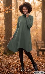 Frau im herbstlichen Wald trägt grünes Baumwoll-Kleid mit langem Arm und Rollkragen, schwarze Strumpfhose und schwarze Schnürstiefel, umgeben von gefallenen Herbstblättern, lächelt freundlich, sitzt auf Holzbank im Hintergrund
