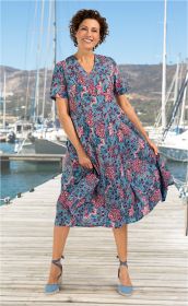 Frau trägt blaues Baumwoll-Kleid mit floralem Muster in Rot und Pink, steht lachend auf Holzsteg mit Segelbooten und Bergen im Hintergrund, trägt hellblaue Espadrilles mit Knöchelband