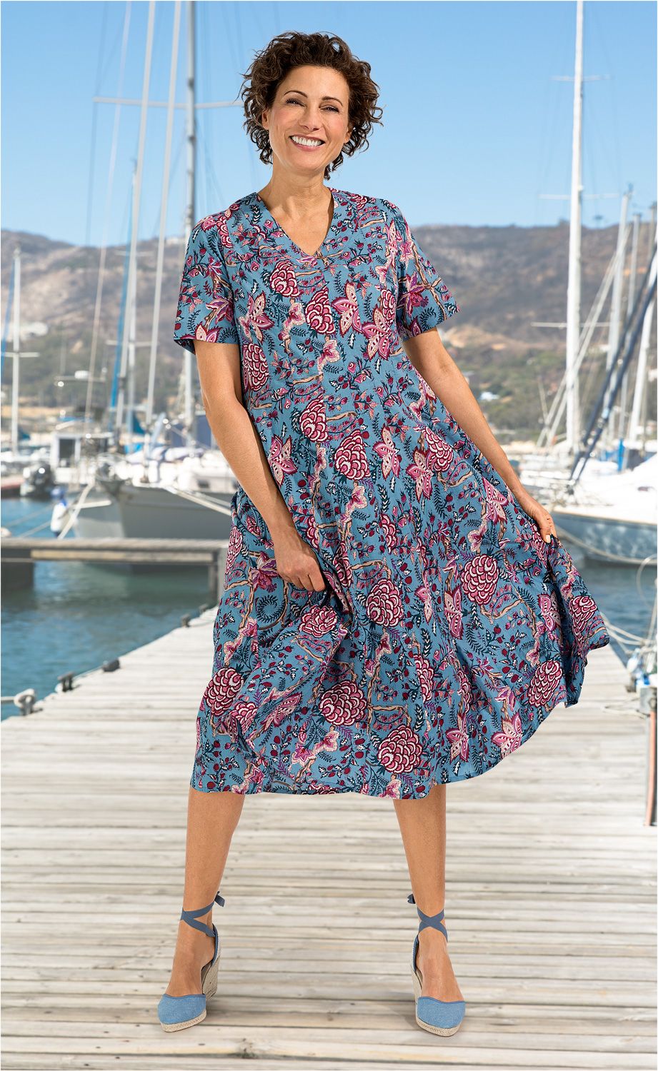 Frau trägt blaues Baumwoll-Kleid mit floralem Muster in Rot und Pink, steht lachend auf Holzsteg mit Segelbooten und Bergen im Hintergrund, trägt hellblaue Espadrilles mit Knöchelband