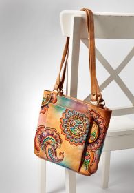 Braune Handtasche mit buntem floralen Muster und Paisley-Design, aus Leder, mit länglichen Henkeln, hängend an weißem Holzstuhl mit Kreuzlehne vor neutralem Hintergrund.