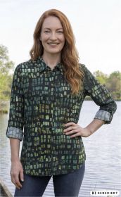 Frau mit roten Haaren trägt langärmlige Baumwoll-Bluse mit grün-blauem Karomuster, Ärmel hochgekrempelt, steht lächelnd vor Gewässer und Bäumen, trägt blaue Jeans