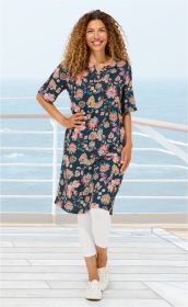 Frau mit lockigem Haar trägt florales Baumwoll-Kleid in Blau mit bunten Blumenmustern, weiße Leggings und weiße Sneaker, steht lächelnd auf einem Holzdeck mit Meeresblick im Hintergrund