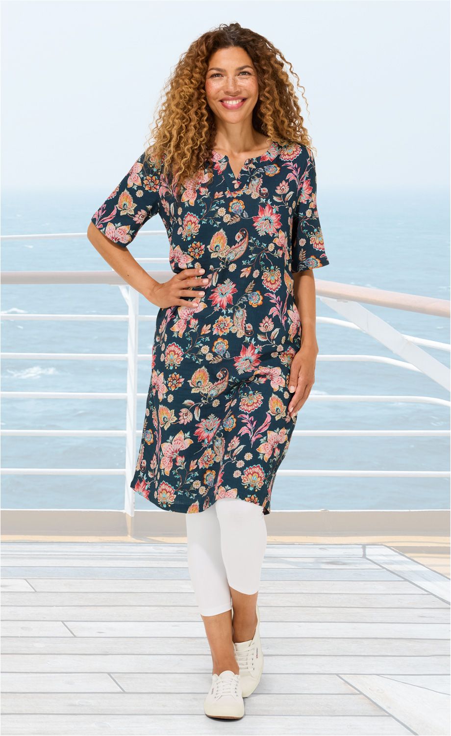 Frau mit lockigem Haar trägt florales Baumwoll-Kleid in Blau mit bunten Blumenmustern, weiße Leggings und weiße Sneaker, steht lächelnd auf einem Holzdeck mit Meeresblick im Hintergrund