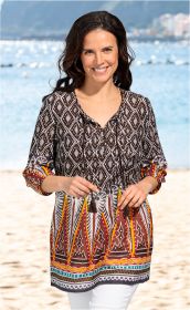 Frau am Strand trägt farbenfrohe Baumwoll-Bluse mit geometrischem Muster in braun, orange, weiß und blau, 3/4-Ärmeln, locker sitzend, dazu weiße Hose, lächelnd, Hintergrund mit Meer und Bergen unscharf