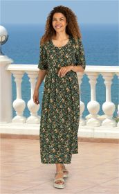 Frau im dunkelgrünen Viskose-Kleid mit buntem Blumenmuster, halblangen Ärmeln und Rundhalsausschnitt, sommerliche Sandalen, steht auf Terrasse mit weißem Geländer und Blick auf das blaue Meer im Hintergrund
