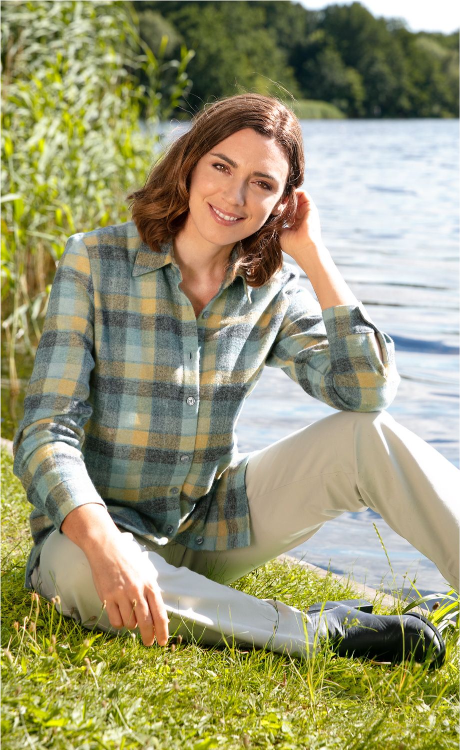 junge Frau mit braunen Haaren und kariertem Hemd sitzt lächelnd auf Gras am Ufer eines Gewässers, im Hintergrund Bäume, sonniger Tag