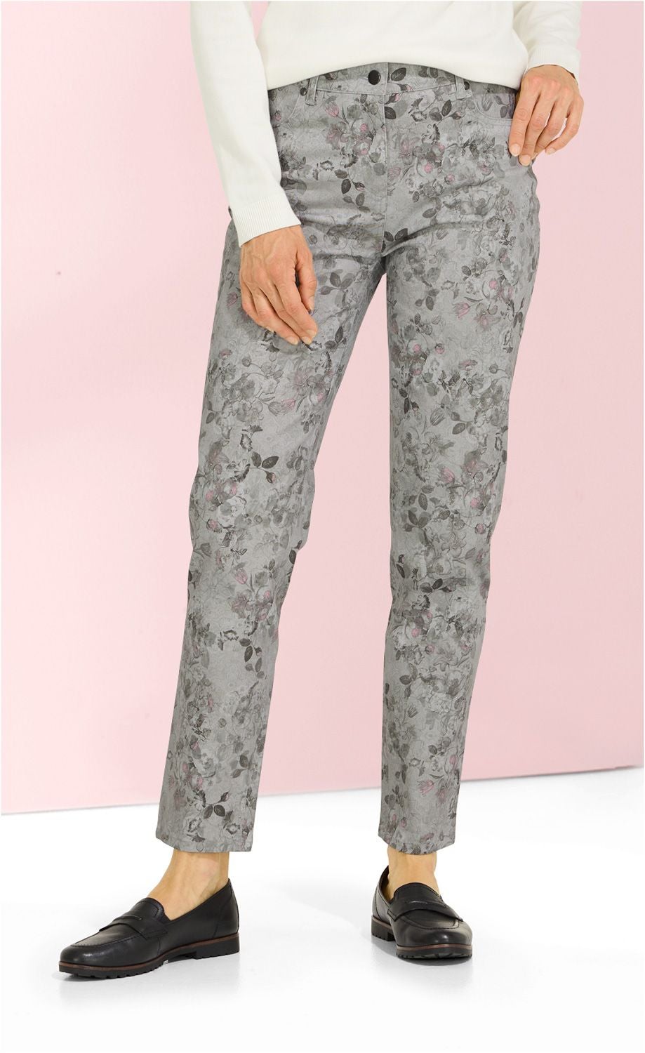 Baumwoll-Mix Hose Damen, hellgrau mit floralem Muster in dunklen und rosa Tönen, gerade geschnitten, mit Hosentaschen, kombiniert mit weißem Oberteil und schwarzen Slippern, vor rosa Hintergrund