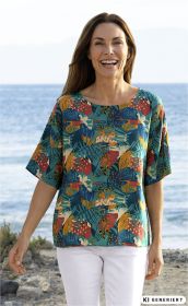 Frau am Strand trägt bunte Viskose-Bluse mit abstraktem Blumen- und Blättermuster in Orange, Blau und Grün, kombiniert mit weißer Hose, lachend und leicht nach vorne schreitend.