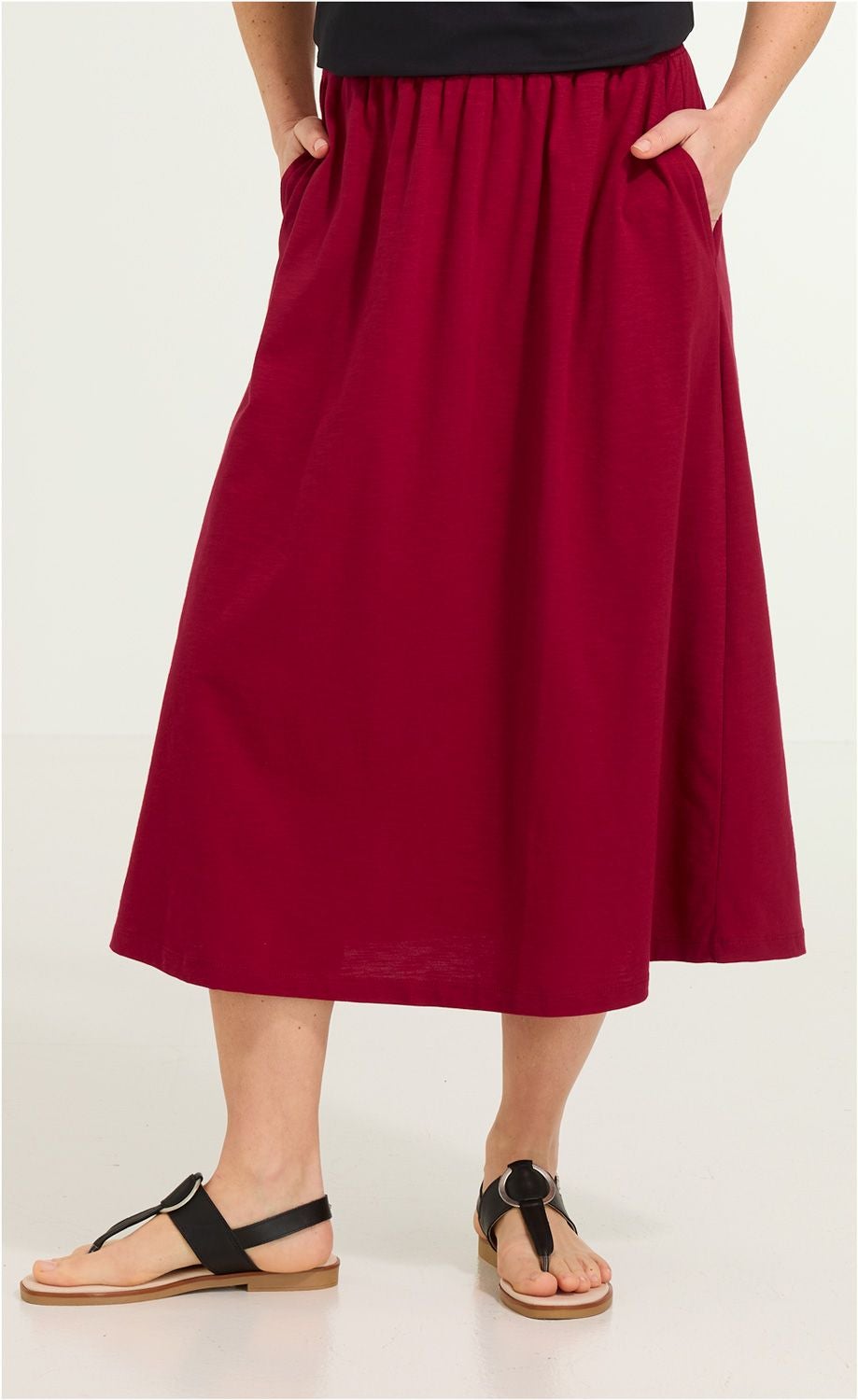 Roter Baumwoll-Midirock mit Gummibund und Taschen, getragen mit schwarzen Sandalen, sichtbar von der Taille bis zu den Füßen, vor hellem Hintergrund