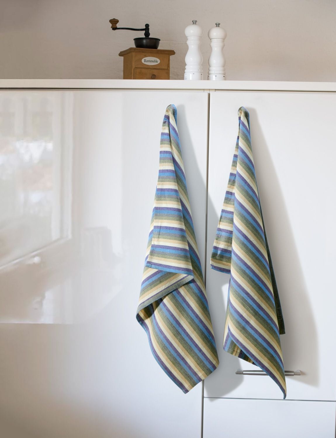 Zwei Baumwoll-Geschirrtücher mit diagonal verlaufenden Streifen in Blau, Grün, Beige und Weiß hängen an weißen Küchenschranktüren, darüber stehen eine alte Holz-Kaffeemühle und zwei weiße Gewürzmühlen.