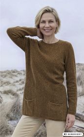 Frau trägt braunen Merino-Mix-Pullover mit langen Ärmeln und zwei Taschen, lächelt, blondes kurzes Haar, helle Stoffhose, steht in einer Dünenlandschaft bei bewölktem Himmel