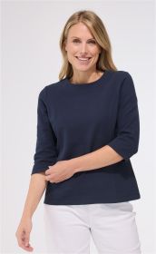 Frau mit blonden Haaren trägt dunkelblaues Baumwoll-Shirt mit 3/4-Ärmeln und rundem Ausschnitt, kombiniert mit weißen Hosen, vor hellem Hintergrund, lächelnd, rechte Hand hält linken Arm.
