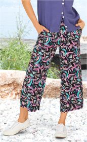 Frau trägt bunte Viskose-Hose mit abstraktem Muster in Schwarz, Lila, Blau, Türkis und Rosa, kombiniert mit weißer Sneaker und lila Strickjacke, steht auf weißem Kiesstrand mit grünem Gebüsch und Wasser im Hintergrund.