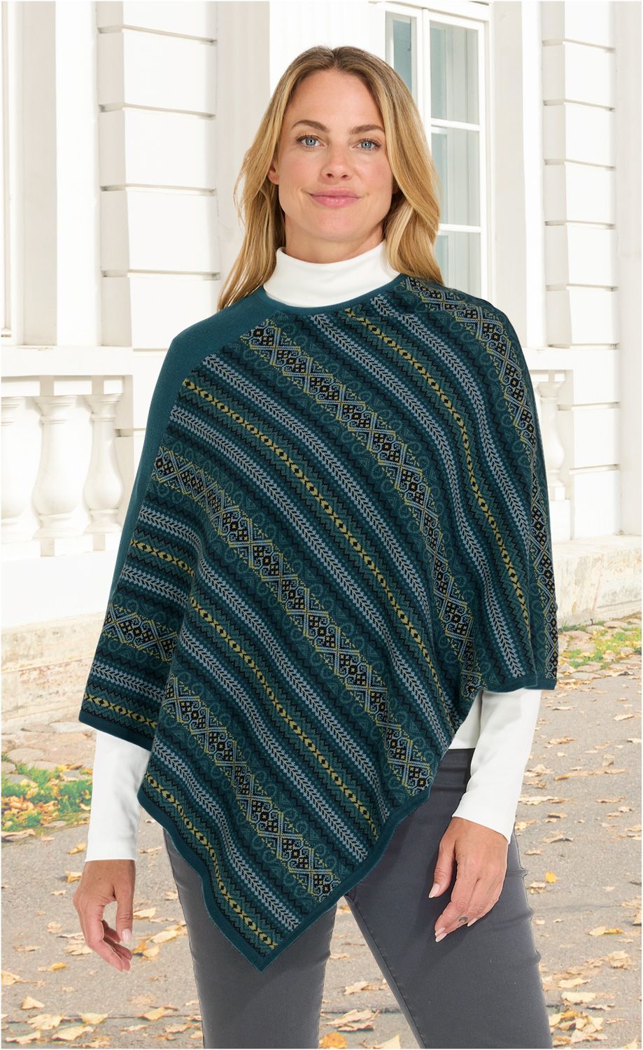 Frau trägt Baumwoll-Mix-Poncho in Blau mit diagonalen Mustern in Oliv, Schwarz und Grau, kombiniert mit weißem Rollkragenpullover und grauer Hose, steht vor weißem Gebäude mit Fenstern und Säulen, Herbstlaub auf dem Boden