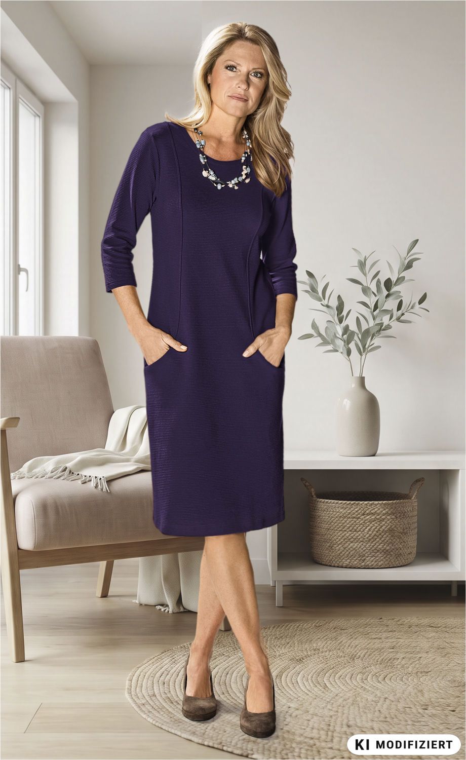 Frau in blauem Baumwoll-Kleid mit 3/4-Ärmeln, stehend im Wohnzimmer, Hände in Taschen, braune Schuhe, blonder mittellanger Haar, Hintergrund mit Sessel, Pflanzenvase und Korb auf hellem Teppich