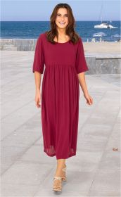 Frau läuft am Strandpromenade, trägt langes, locker fallendes, dunkelrotes Viskose-Kleid mit halblangen Ärmeln und Sandalen, im Hintergrund Meer, Segelboot und Strand mit Sonnenschirmen sichtbar