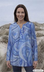 Frau mit langen braunen Haaren trägt blaue Viskose-Tunika mit abstraktem Muster und Knopfleiste, steht vor Dünenlandschaft mit Gras, lächelnd, natürliche Beleuchtung