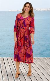 Frau steht auf Holzsteg am Meer trägt buntes Viskose-Kleid in Rot, Lila und Orange mit dreiviertel Ärmeln, V-Ausschnitt, knieumspielender Länge, trägt braune Sandalen, lächelt, Hintergrund Küstenlinie und Wasser bei klarem Himmel