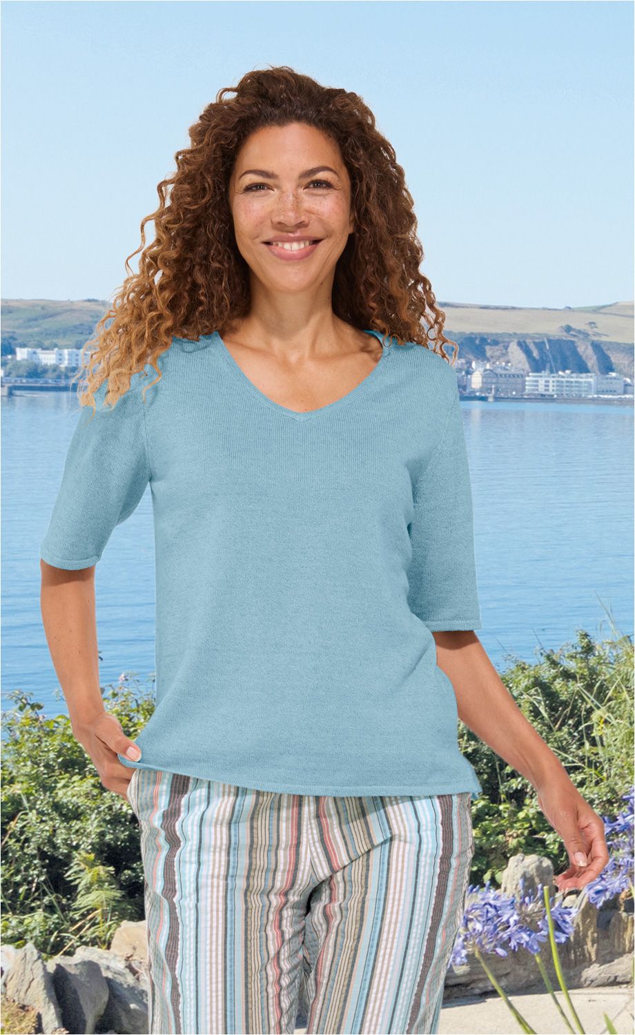 Frau mit lockigen braunen Haaren trägt hellblauen Leinen-Mix-Pullover mit V-Ausschnitt und halblangen Ärmeln, stehender vor einem See mit grüner Vegetation und blühenden Pflanzen im Sommer, gestreifte sommerliche Hose