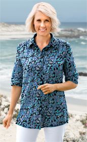 Frau mit kurzen blonden Haaren trägt Viskose-Mix Bluse mit blau-türkis floralem Muster, halblange Ärmel, Knopfleiste, vor sandigem Strand mit Meer im Hintergrund, weiße Hose