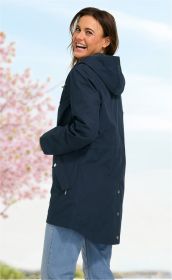 Frau mit dunkler Baumwoll-Jacke mit Kapuze, seitlich stehend, lachend, vor unscharfem Frühlingshintergrund mit blühendem Baum und blauem Himmel, trägt helle Jeans