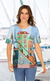 Frau trägt hellblaues Baumwoll-Shirt mit buntem Druck von Häusern, Treppe, Fahrrädern und Katzen, dazu blaue Jeans, Hintergrund unscharf mit Yachten und Steg