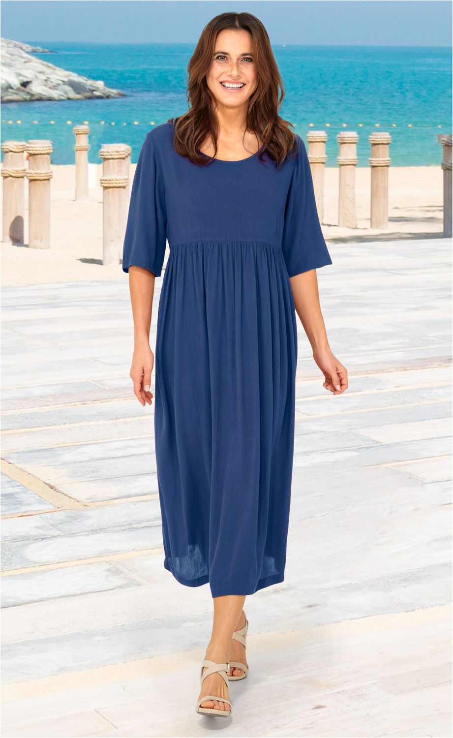 Frau am Strand trägt ein knielanges, blaues Viskose-Kleid mit halblangen Ärmeln und beigen Sandaletten, sonniger Tag mit Meer und Felsen im Hintergrund