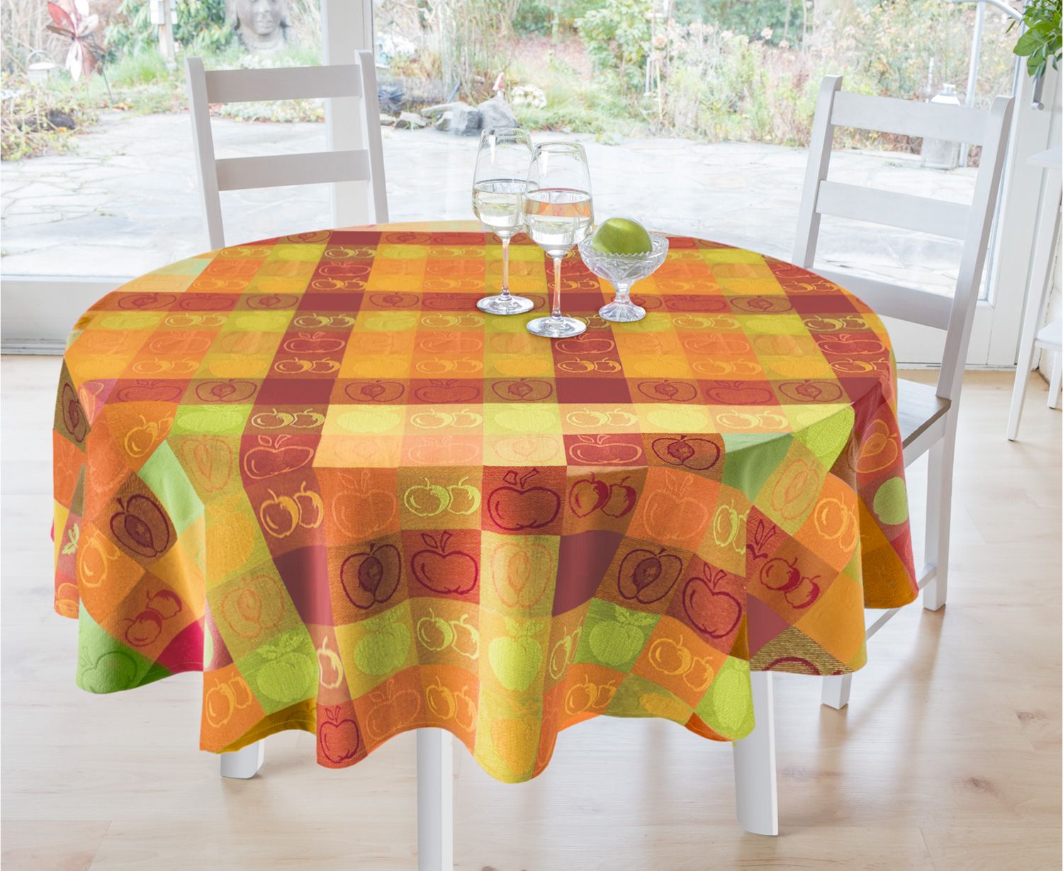runde Tischdecke 130x180 cm mit kariertem Muster in Orange, Rot, Gelb und Grün, dekoriert mit Apfelmotiven, auf Holztisch mit zwei weißen Stühlen, darüber zwei gefüllte Weingläser und eine Glasschale mit grünem Apfel, heller Holzfußboden und Gartenansicht