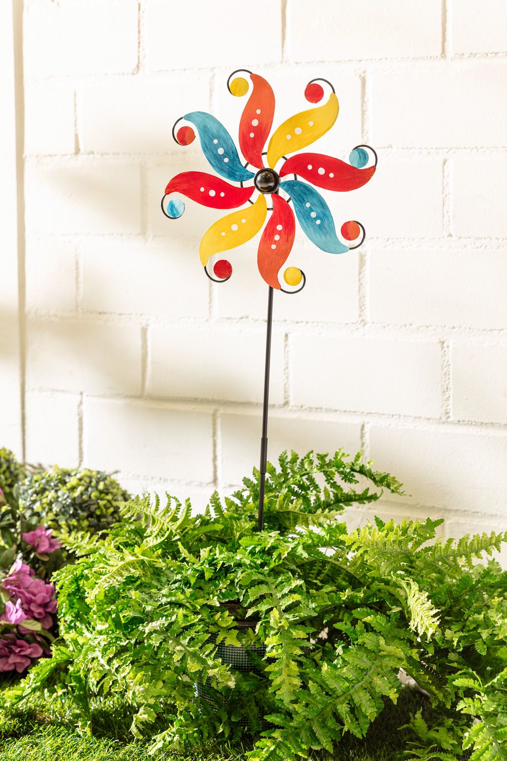 bunter Deko-Gartenstecker mit rot, gelb und blau lackierten, gebogenen Metallblättern auf schwarzem Stab, eingesetzt in grünem Farn vor weißer Mauer mit rosa Blüten im Hintergrund